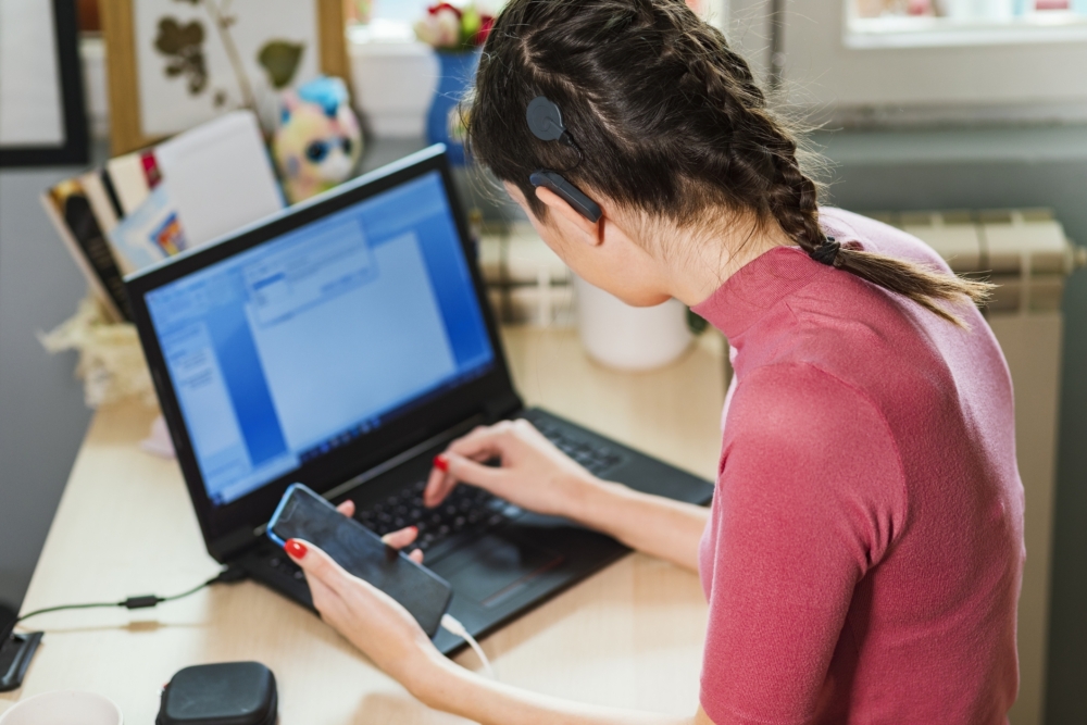 A woman with a cochlear implant sits at a desk holding a phone with one hand and typing on a laptop with the other