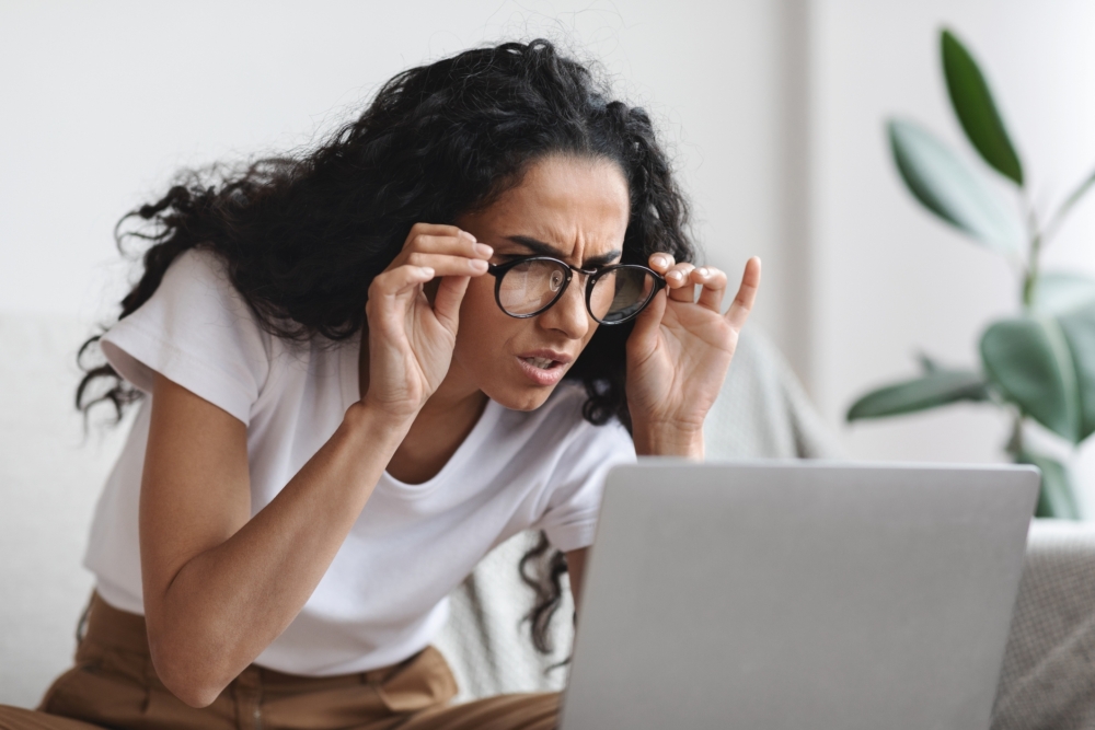 A woman holding her glasses and leaning forward to squint at her laptop screen