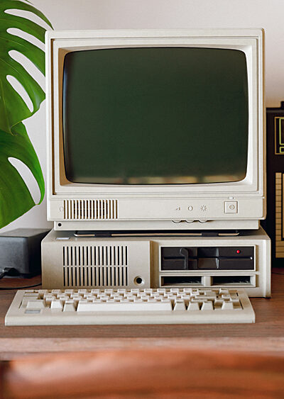Retro computer stands on a desk with a plant, keyboard, computer mouse, retro radio, and phone.