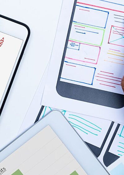 A phone displaying an accessible website next to a hand holding a pen, website designs on paper, and the corner of a tablet