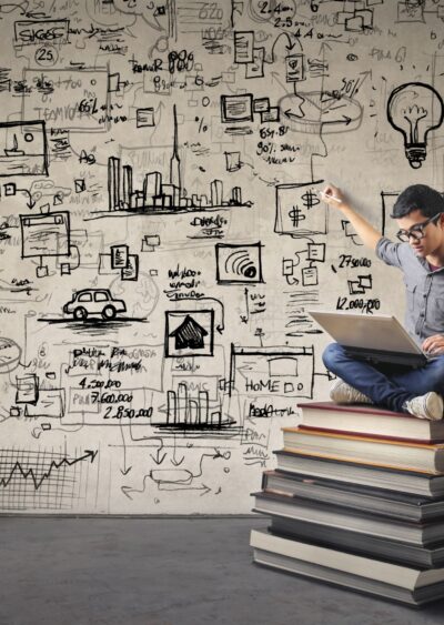 Student sitting on a stack of book with a laptop making graphics on wall